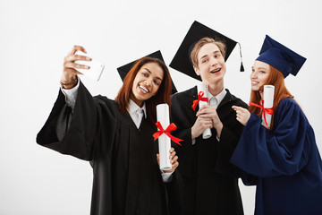 Three international graduate friends rejoicing in mantles making a selfie on a phone. Future lawyers or doctors or engineers having fun with diplomas. White background.