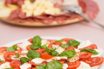 Tomato and mozzarella salad with basil leaves, sprinkled with pepper, olive oil and acceto balsamico on the white plate; with salami and cheese tray in the background