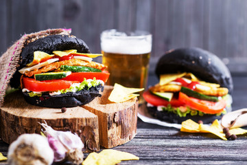 Black burger with chicken, lettuce, mayonnaise, ketchup, tomato, cucumber, cheese, corn chips and a light beer on a dark wooden background, a roll with cuttlefish ink 