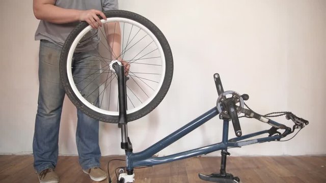 A blond bearded man in a blue T-shirt and jeans fastens the front wheel to the mountainbike frame in the room next to the white wall.