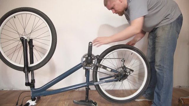 A blond bearded man in a blue T-shirt and jeans fastens the rear wheel to the mountainbike frame in the room next to the white wall.