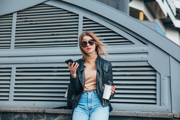 Fototapeta premium Fashion portrait pretty woman in black rock style in sunglasses using mobile phone over gray background in city .Girl with cofee