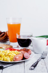 Hearty and traditional breakfast, scrambled eggs and milk with grilled pork, vegetables, tomato, cucumber, toast, juice and black coffee on a white wooden background