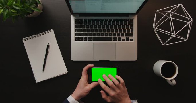 Business man using smart phone, checking information on cell phone with green screen on brown background with laptop. Office desk background. Slow motion. Top view. Chroma key. Horizontal position.