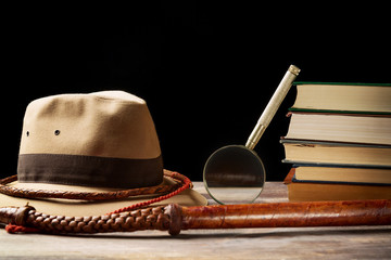 Fedora hat with bullwhip near magnifying glass and old books on black background. Adventure concept - obrazy, fototapety, plakaty