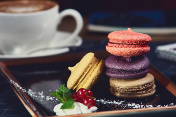 Colorful macaroons served on black stylish plate and cup of cappuccino coffee at background