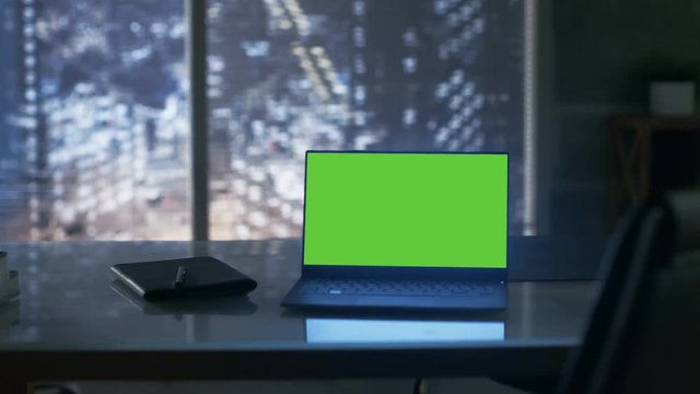Close-up Footage of a Laptop with Green Mock-up Screen Standing on a Wooden Table. In the Background Big City Window View.Shot on RED EPIC-W 8K Helium Cinema Camera.