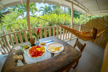 Table with breakfast and fruits and woman relaxing in the hammock in a terrace with tropical garden view
