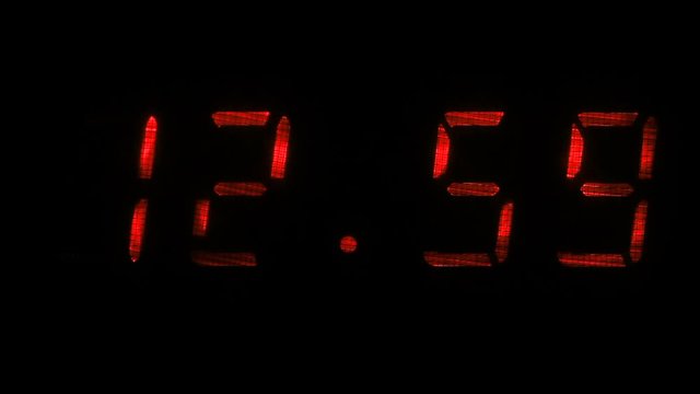 Digital clock with fluorescent display shows the time of 12 hours 59 minutes to 13 hours 00 minutes in the red on a black background