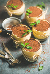 Homemade Italian dessert Tiramisu served in individual glasses with mint leaves and cocoa powder over grey concrete background, selective focus, vertical composition
