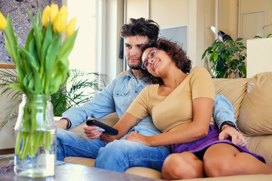 The attractive casual couple sits on a couch and switching tv channels with remote control.