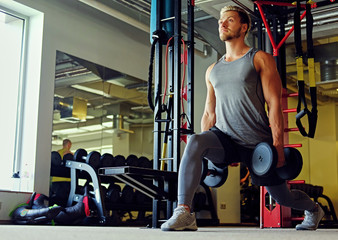 Portrait of a blond sporty male doing squats with dumbbells in a gym club.