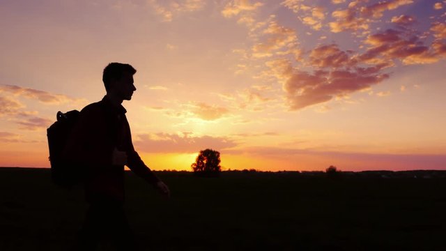 A teenager with a backpack over his shoulder goes towards the sunset in the field or in the countryside. Silhouette video, side view. Concept - new research, forward to the unknown, to leave home