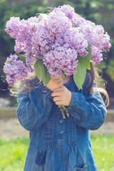 Little girl holding lilac flowers in front of her face in the garden, toned photo