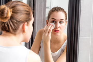 young woman removing makeup with swab in front of mirror