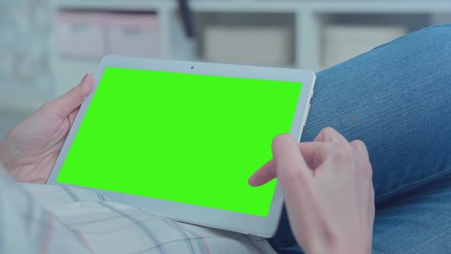 Young Woman in blue jeans laying on couch uses Tablet PC with pre-keyed green screen. Few types of gestures - scrolling up and down, tapping, zoom in and out. Perfect for screen compositing.