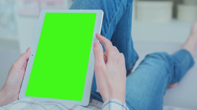 Young Woman in blue jeans laying on couch uses Tablet PC with pre-keyed green screen. Few types of gestures - scrolling up and down, tapping, zoom in and out. Perfect for screen compositing. 