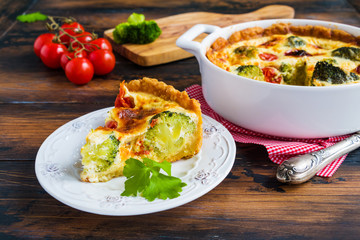 Homemade quiche with broccoli and cherry tomatoes in the baking dish and a piece of cake on a plate on the rustic wooden table.