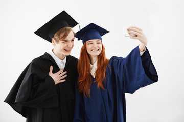 Two graduate friends in caps and mantles laughing making selfie over white background before receiving their magister diploma or bachelor of arts or other academic degree. Study concept.