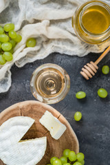 French soft cheese from Normandy region sliced on a wooden cut with green grapes, honey, spoon and glass of white wine on dark rustic table, blurred background, top view