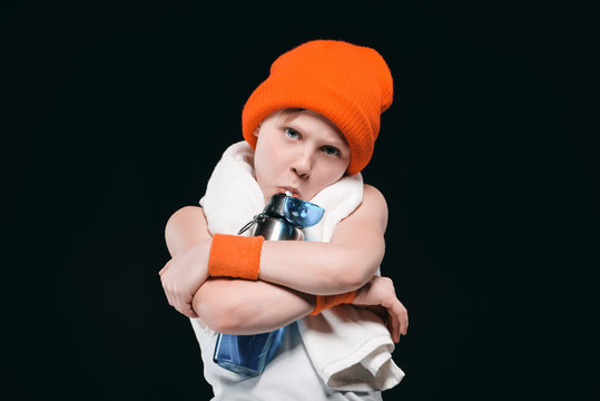 Boy in hat with towel on neck hugging bottle and drinking water isolated on black