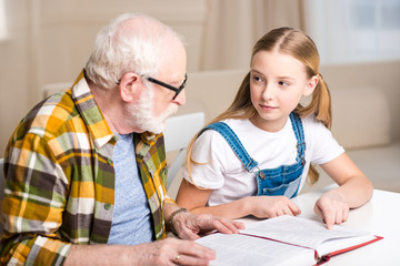 Grandfather and granddaughter sitting at table and reading book together