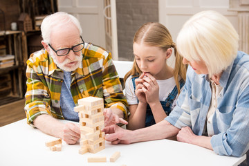 Concentrated little girl with grandparents playing jenga game together at home