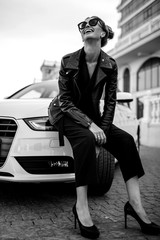 fashion outdoor photo of sexy beautiful woman with dark hair in black leather jacket and sunglasses posing in luxurious auto