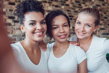 Beautiful and funny girls in white T-shirts makes selfie. Two afro-americans and one european.