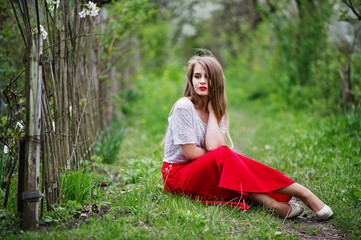 Portrait of sitting beautiful girl with red lips at spring blossom garden on green grass, wear on red dress and white blouse.