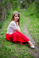 Portrait of sitting beautiful girl with red lips at spring blossom garden on green grass, wear on red dress and white blouse.