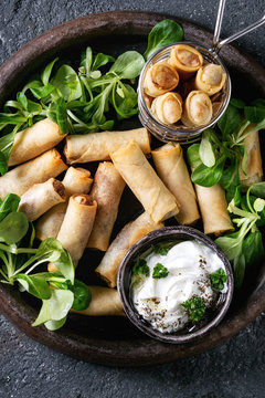 Fried spring rolls with white yogurt sauces, served in terracotta plate and fry basket with fresh green salad over black texture background. Flat lay. Asian food