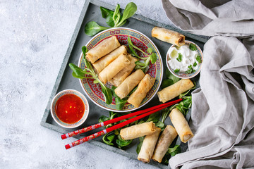 Fried spring rolls with red and white sauces, served in china plate on wood tray with fresh green salad and wooden chopsticks over gray blue texture background. Flat lay, space. Asian food