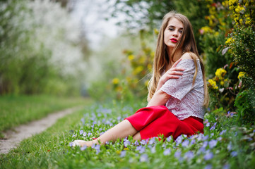Naklejka na ściany i meble Portrait of sitiing beautiful girl with red lips at spring blossom garden on grass with flowers, wear on red dress and white blouse.