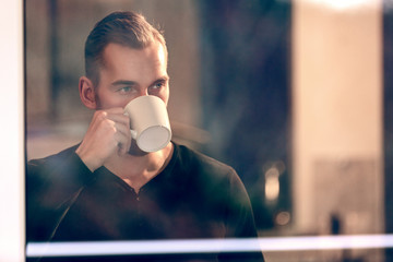 Handsome man drinking from a mug looking out of the window from inside house, wearing a black shirt.