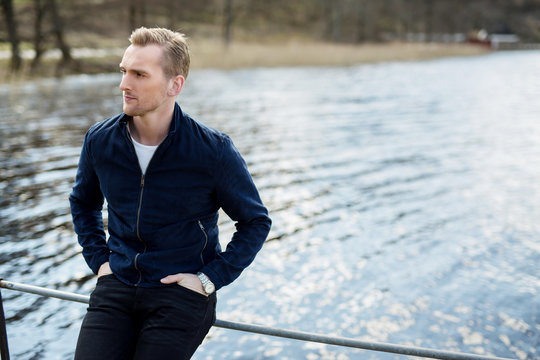 Attractive Pensive Man Wearing A Blue Leather Jacket Sitting Down Outdoors On A Sunny Day With The Lake Behind Him, Looking Away From Camera.