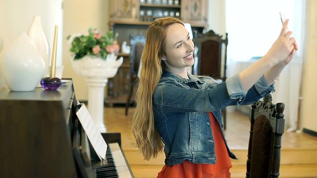 Happy girl sitting next to the piano and doing selfies on smartphone, steadycam shot
