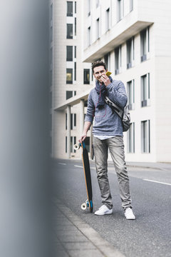Businessman carrying skateboard, using smartphone and earphones