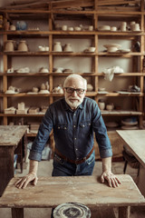 Front view of senior potter standing and leaning on table against shelves with pottery goods at workshop