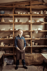Fototapeta na wymiar Front view of senior potter in apron standing against shelves with pottery goods at workshop