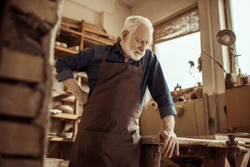 Front view of senior potter in apron standing and leaning on table against shelves with pottery goods at workshop