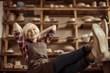 Naklejka premium Front view of senior woman sitting on chair with legs on table against shelves with pottery goods