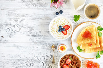 Overhead of traditional russian breakfast on white wooden table with copy space. Pancakes with caviar, cottage cheese with strawberry and blueberry, cornflakes, boiled egg and coffee.