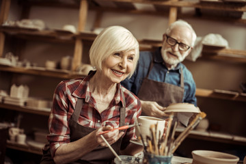 Woman painting clay pot with senior potter at workshop