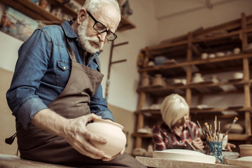 Senior potter in apron and eyeglasses examining ceramic bowl with woman working on background