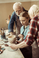 Side view of girl painting clay pot and grandparents helping at workshop