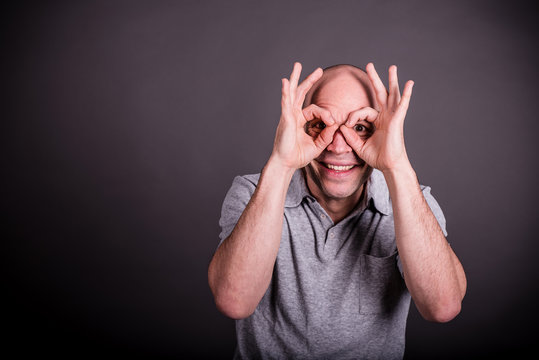 Mann mit grauem Poloshirt formt lachend mit seinen Fingern eine Brille, lacht dabei