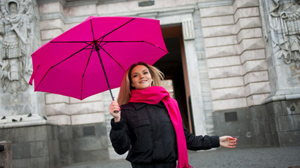Beautiful young and happy blond woman with colorful umbrella on the street. The concept of positivity and optimism. Girl in a bright pink scarf and umbrella walking in a rainy city.