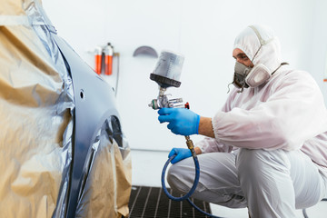 Man with protective clothes and mask painting car using spray compressor. Selective focus. 