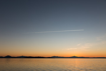 A dusk at a lake, with a long contrail in the middle of an almost clear sky